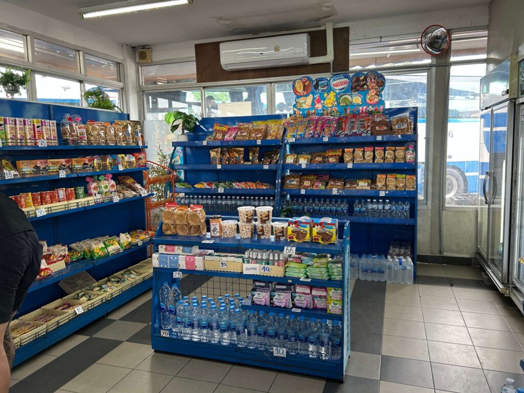 Interior view of a bus terminal shop in North Pattaya, showing shelves stocked with bottled water and packaged snacks
