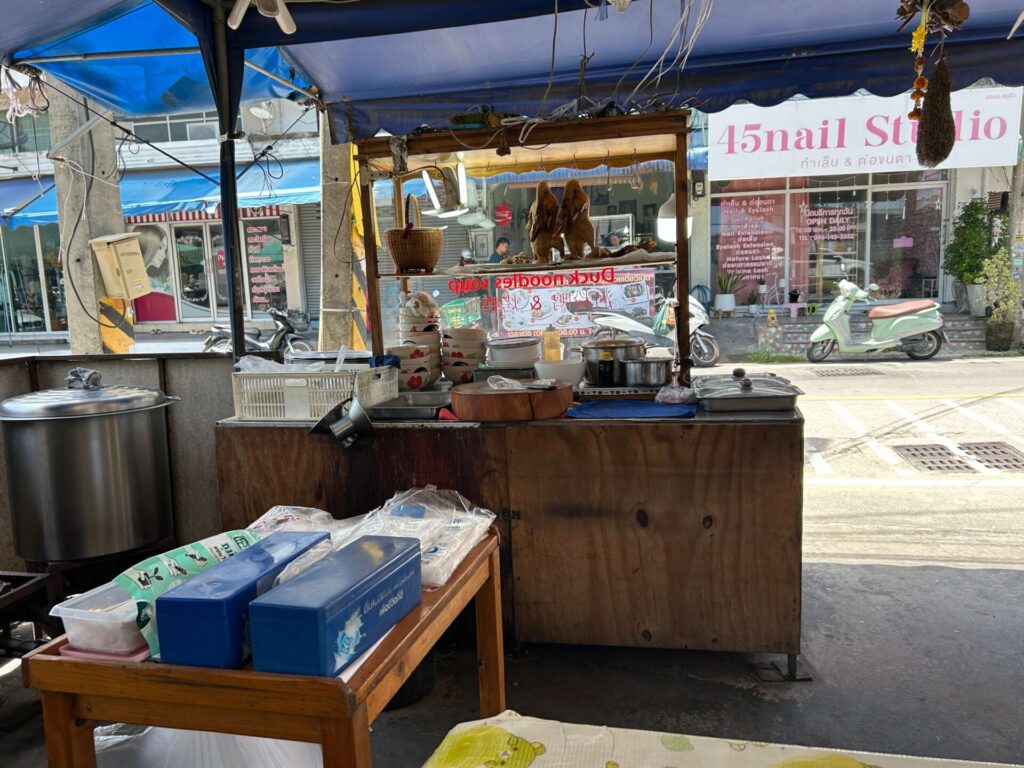 Cooking area of a street food stall with hanging roasted ducks and various ingredients prepared for making noodles