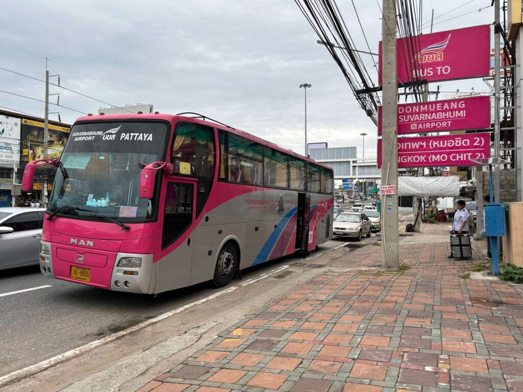 Pink and grey Bor Kor Sor bus parked on the roadside with a sign indicating service to Suvarnabhumi Airport and Pattaya