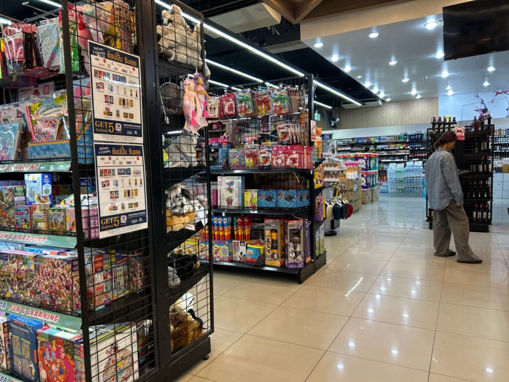 An aisle within Villa Market at Little Walk Pattaya, showing shelves packed with a diverse range of items including toys, stationery, and other miscellaneous goods, providing convenient options for bus travelers