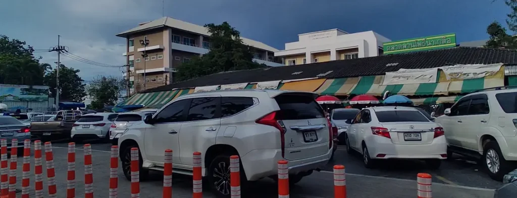 The entrance to Lan Pho Naklua Market, showing rows of cars parked and a building with striped awnings in the background. The scene suggests a busy, bustling market day.