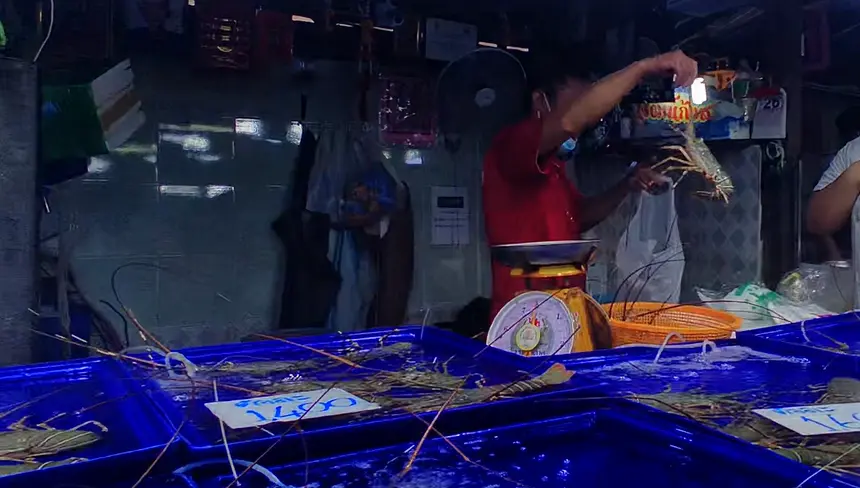 A stall at the Naklua seafood market where a vendor is holding up a fresh lobster. Other fresh seafood is displayed in blue plastic containers with prices clearly visible on signs.