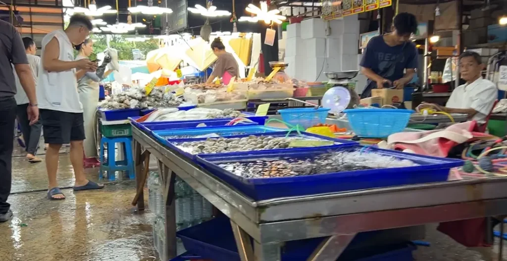 A seafood stall at Lan Pho Naklua Market in Pattaya at night. The stall is lit up by bright lights, and several people are seen looking at the fresh seafood laid out on display.