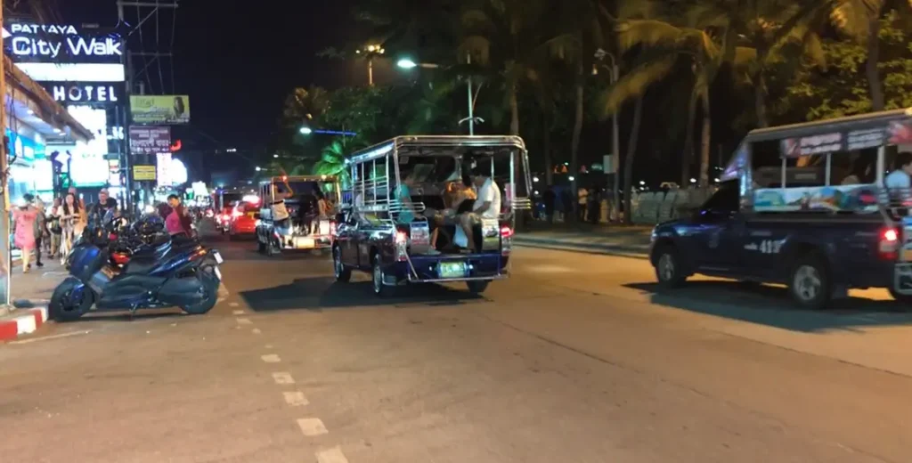 A night shot of a songthaew driving along Pattaya's Beach Road. The street is illuminated by streetlights, and a scooter is parked on the left side of the road with a hotel sign in the background.
