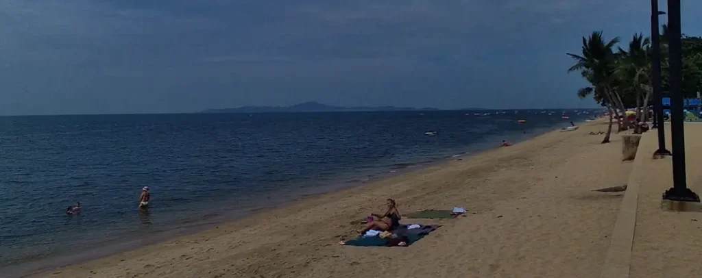 A tranquil view of Jomtien Beach in Pattaya on a cloudy day, showing beach chairs and umbrellas under the shade of palm trees along the sand.