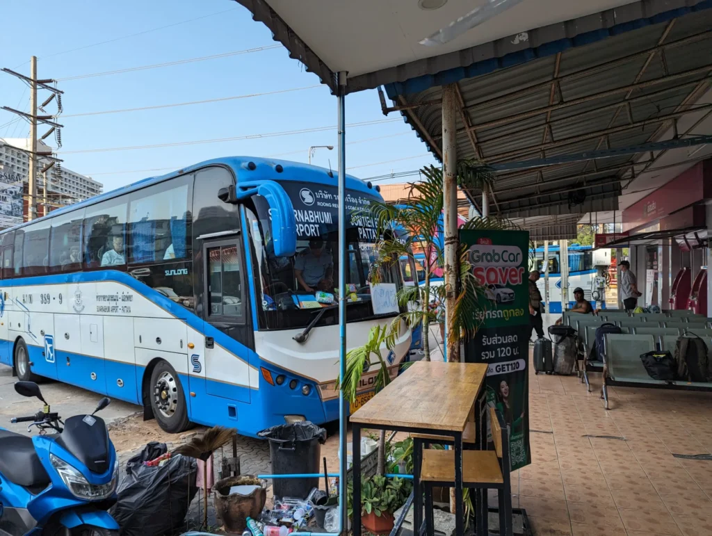 An image of the Pattaya Jomtien bus terminal, showing the blue bus to Suvarnabhumi Airport (BKK) waiting in the station. The shot captures a typical bus stop scene with a few people waiting and parked motorcycles.