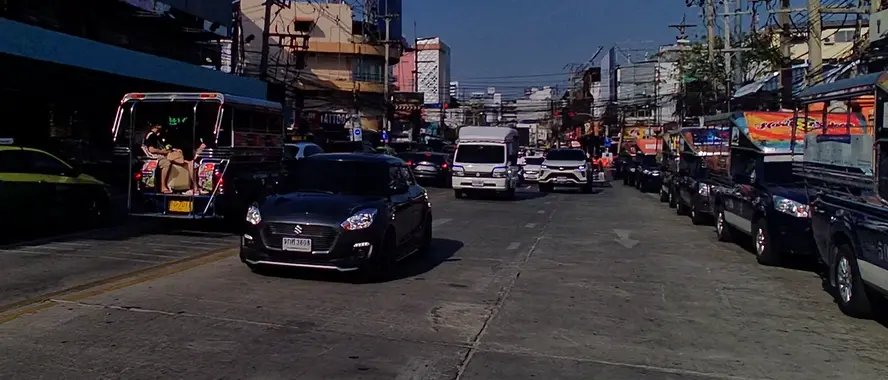A blue songthaew waiting on the right side of the street on the route from Pattaya to Jomtien Beach. A Suzuki Swift and other vehicles are seen driving in the opposite direction towards central Pattaya.