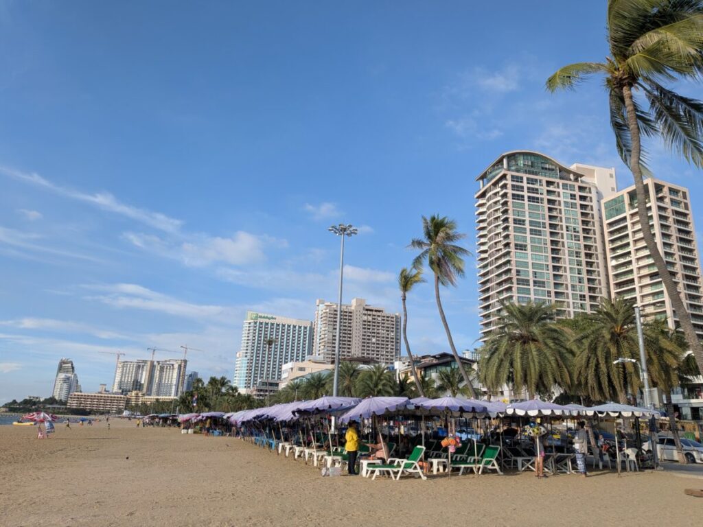 The daytime view of Pattaya Beach Road hotel area, showing tall buildings, beach umbrellas, and the calm blue ocean.