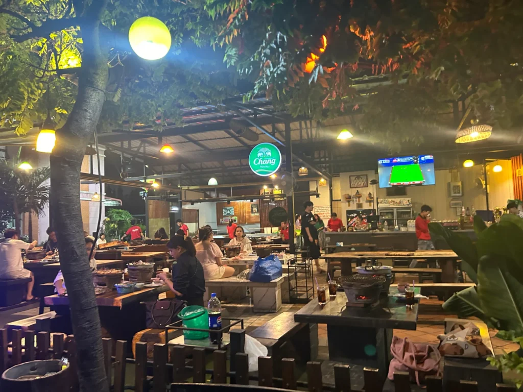Warmly lit, open-air Mookata restaurant interior at night in Pattaya, showing wooden tables, charcoal grills, customers dining, and a Chang beer sign.