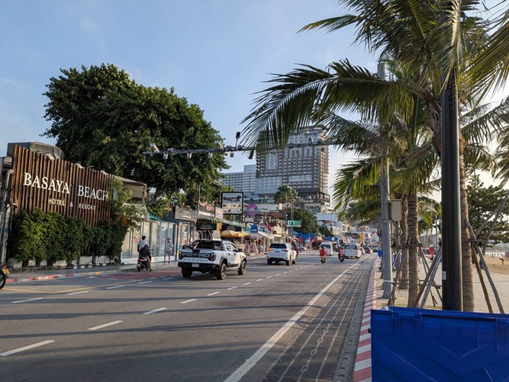 The daytime view of Pattaya Beach Road, showing the calm blue ocean, a few boats, and the skyline of tall hotel buildings along the coast.