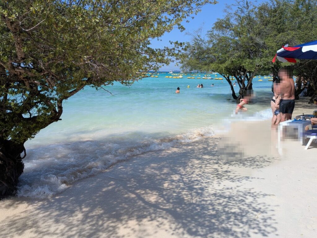 Shaded area at Tien Beach with trees along the water, gentle waves, and visitors relaxing on sunbeds.