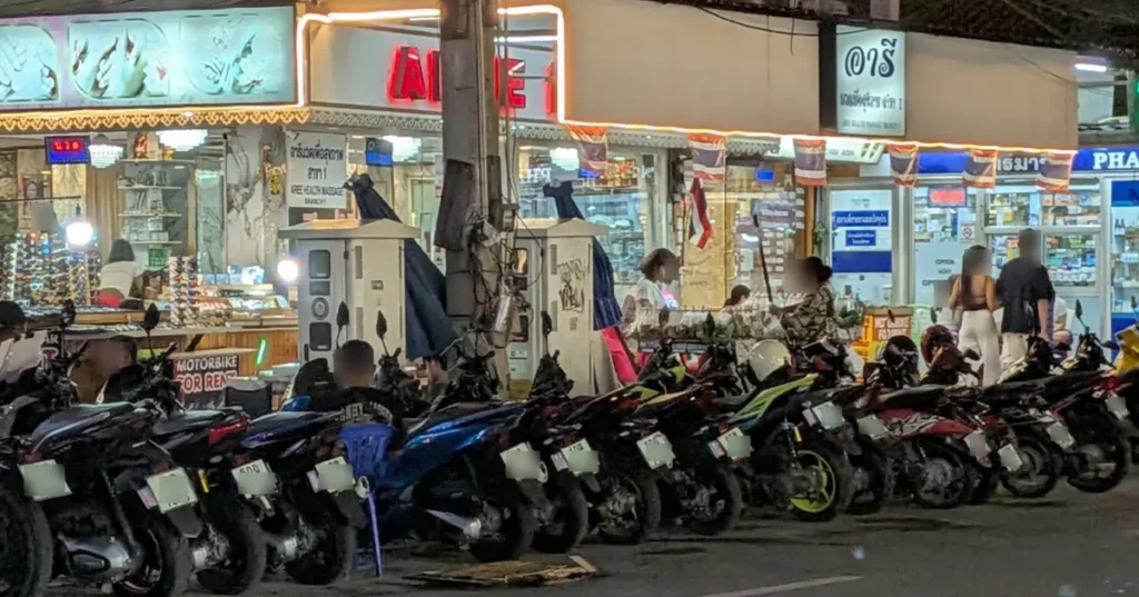 A row of motorbikes parked for rent along a busy street with bright signs at night in Pattaya.