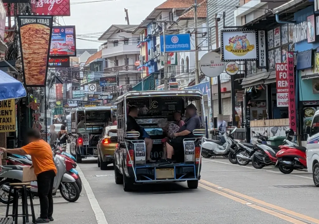 A lively daytime street view of Soi Buakhao in Pattaya with a blue songthaew, local shops, and motorbikes.