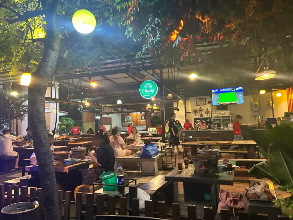 Warmly lit, open-air Mookata restaurant interior at night in Pattaya, showing wooden tables, charcoal grills, customers dining, and a Chang beer sign.